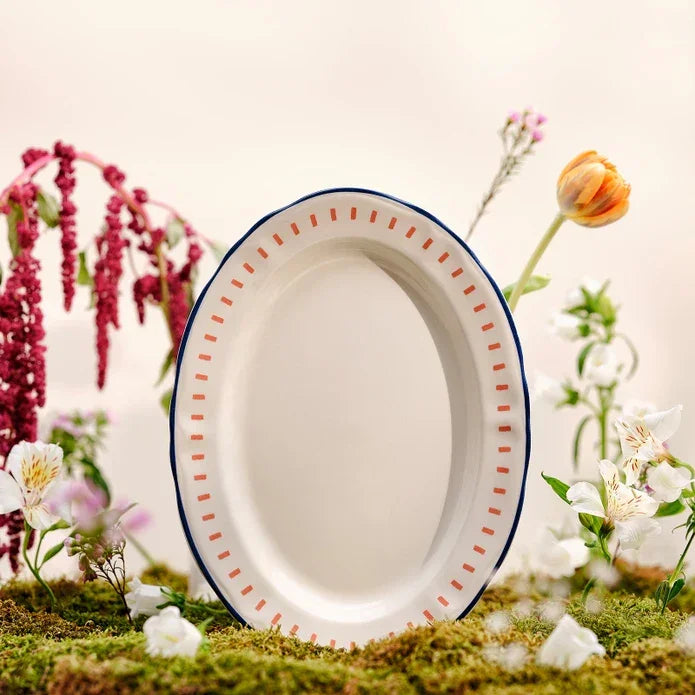 Oval white ceramic dinner plate with orange dotted border and blue rim, surrounded by flowers on moss