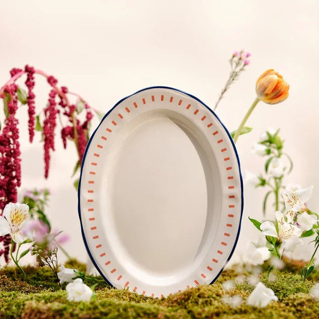 Oval white ceramic dinner plate with orange dotted border and blue rim, surrounded by flowers on moss