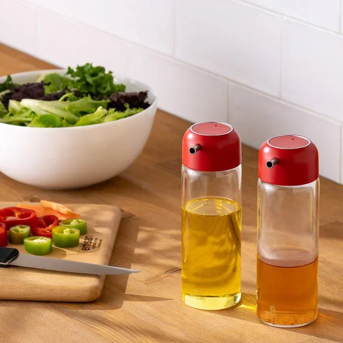 Emsan Jordan glass oil and vinegar dispensers with red tops on wooden kitchen countertop next to fresh salad and sliced peppers